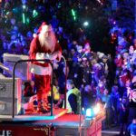 Santa Claus greets a cheering crowd from a fire truck during a nighttime holiday parade filled with lights and festive decorations, capturing the joyful Pop Culture spirit of Christmas celebrations.