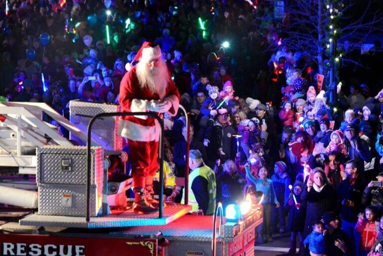 Santa Claus greets a cheering crowd from a fire truck during a nighttime holiday parade filled with lights and festive decorations, capturing the joyful Pop Culture spirit of Christmas celebrations.
