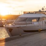 Luxury yacht docked at a marina during sunset, with golden sunlight reflecting on the water and pier in the foreground.