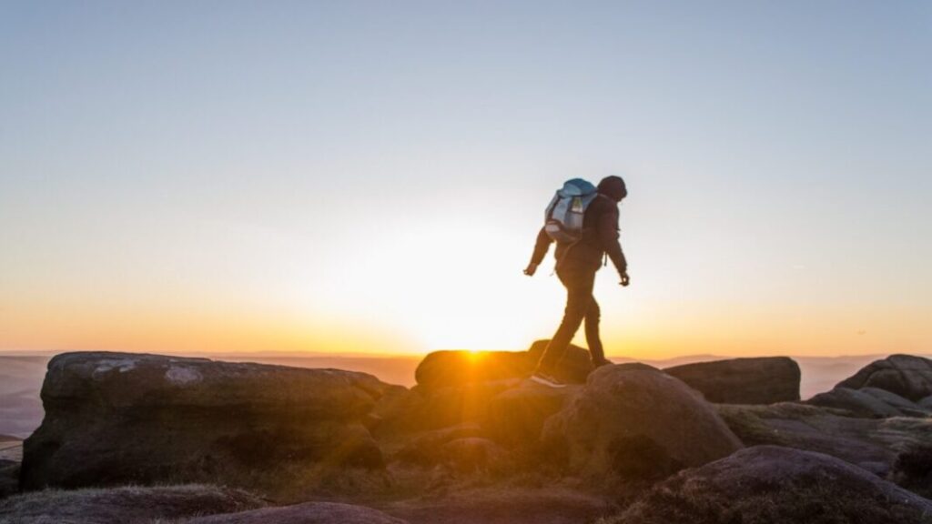 Hiker walking across rocky terrain during sunrise with backpack and scenic mountain landscape in the background.