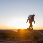 Hiker walking across rocky terrain during sunrise with backpack and scenic mountain landscape in the background.