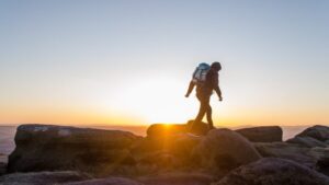 Hiker walking across rocky terrain during sunrise with backpack and scenic mountain landscape in the background.