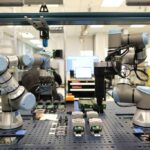 Dual robotic arms operating in an automated manufacturing workstation, handling electronic components on a perforated workbench inside a laboratory or industrial automation environment.
