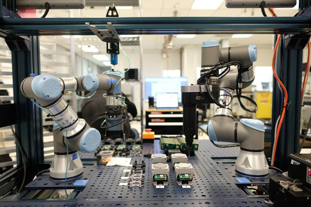 Dual robotic arms operating in an automated manufacturing workstation, handling electronic components on a perforated workbench inside a laboratory or industrial automation environment.