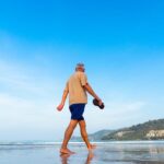 Senior man walking barefoot along a peaceful beach shoreline under a clear blue sky with coastal hills in the background.