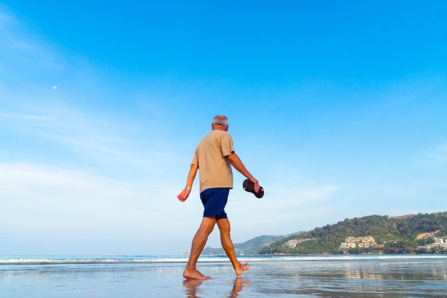Senior man walking barefoot along a peaceful beach shoreline under a clear blue sky with coastal hills in the background.
