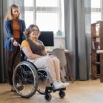 young-smiling-woman-wheelchair-looking-forwards-while-her-sister-friend-standing-helping-her-with-moving-around_274679-17610
