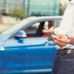 Male hand holding car keys offering new blue car on background