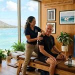 A male patient receives care from a female chiropractor at a light-filled chiropractic care in Honolulu clinic with ocean views and natural wood decor.
