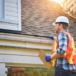 A man with hard hat standing on steps inspecting house roof
