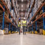 workers-with-hardhats-reflective-jackets-walking-through-big-warehouse-aisle-checking-goods-condition