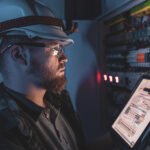 Electrician in overalls, focused on work in switchboard with fuses, using tablet.